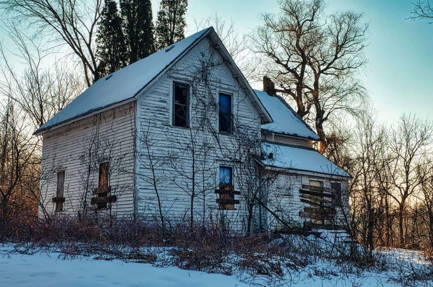 Boarded-up vacant farmhouse surrounded by bare trees in winter